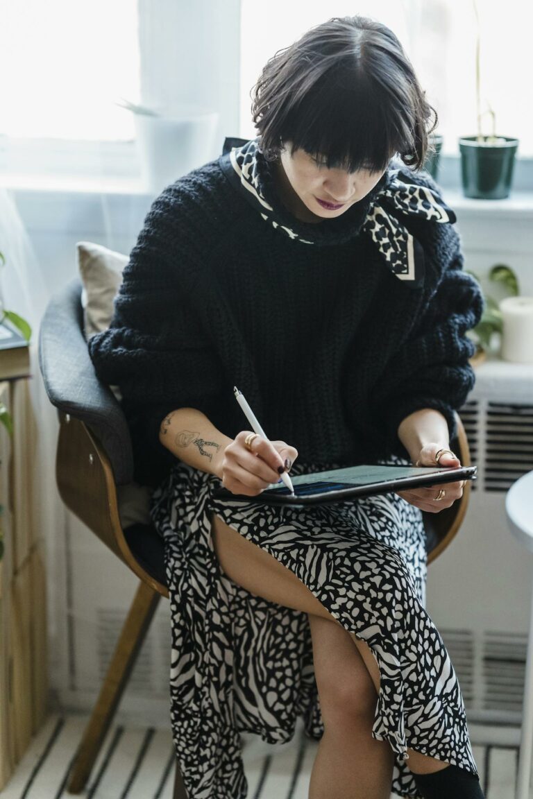 Concentrated young female illustrator in stylish outfit sitting on chair and drawing on graphic tablet in light room against window in daytime